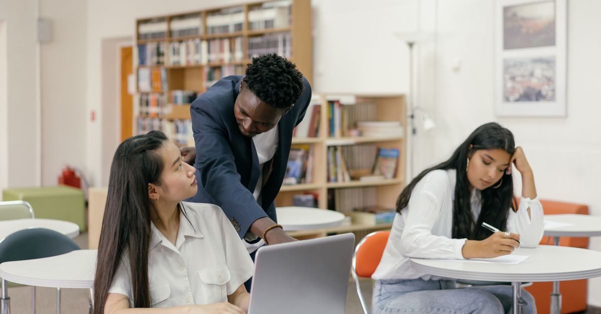 An instructor pointing at a student's laptop screen while providing guidance in a classroom.