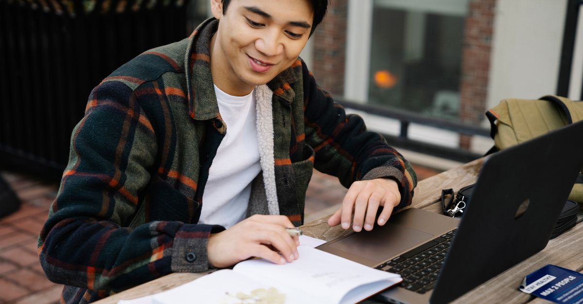 A man sitting at an outdoor wooden table working on a laptop with an open notebook nearby.