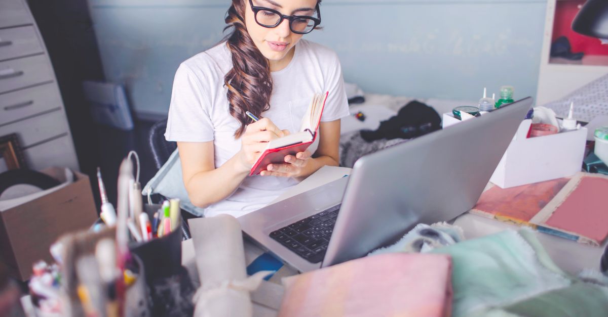 A young woman wearing glasses writing notes in a notepad beside an open laptop.