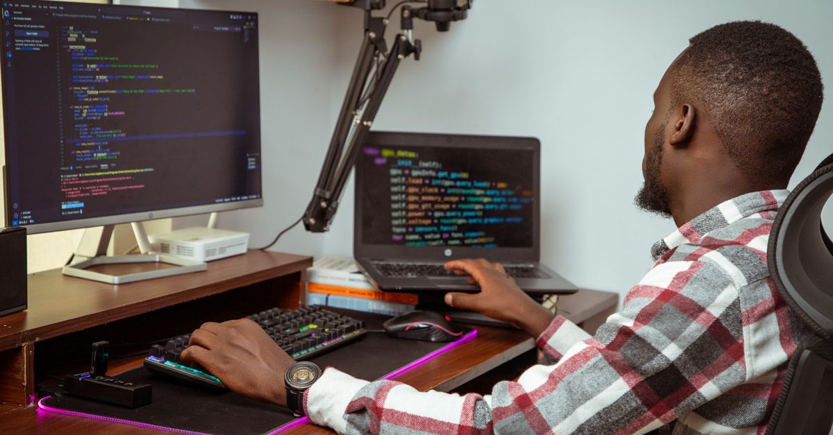 A man sitting at an office desk working on a desktop computer and laptop.