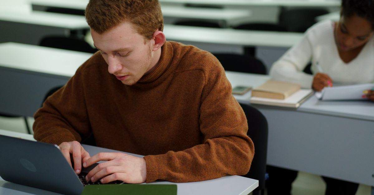 A student in a brown sweater typing on a laptop while working alongside a classmate in a classroom setting.