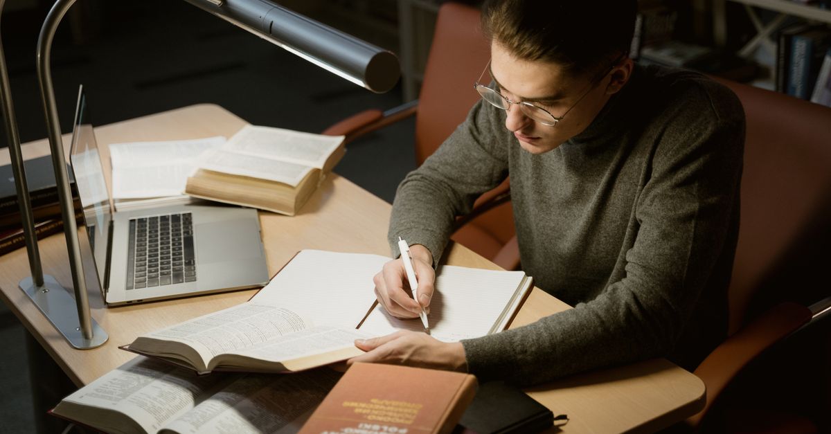 A young adult taking notes at a desk with books and a laptop under a desk lamp.