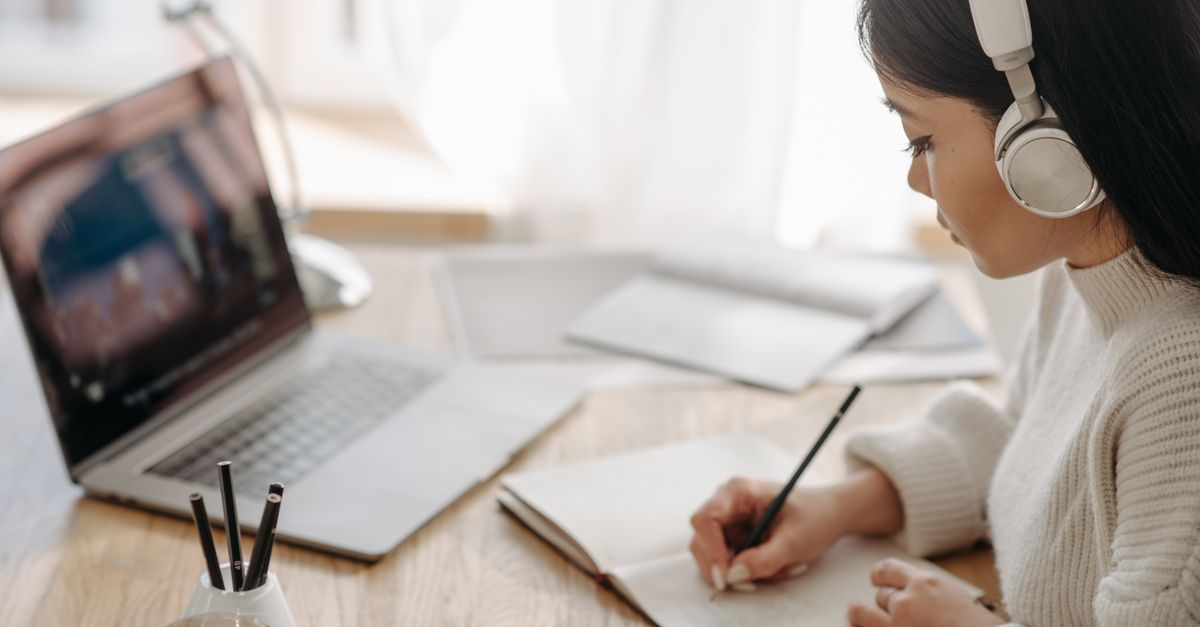 A woman wearing headphones taking notes on paper while following an online study session on a laptop.